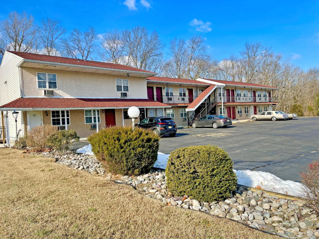 Two story building with exterior room entrances, covered walkways and stairs, parking spaces, grassy areas, landscaping with decorative stones amd small shrubs