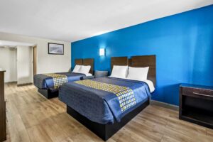 Two queen beds, art image, easy chair, wall bedside light, night stand, laminate flooring, doorway to bathroom