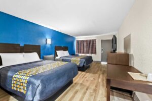 Two queen beds, easy chair, wall bedside light, wooden unit with TV, desk, laminate flooring