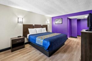 King bed, night stand with telephone, wall bedside lights, art image, wooden unit with TV, doorway to bathroom, laminate flooring