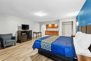 King bed, night stand, wall bedside light, mirror, alcove with kitchenette, occasional table, wooden unit with fridge, microwave, drawers and TV, easy chair, laminate flooring