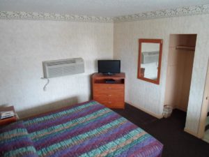 Queen bed, night stand and telephone, wooden drawers unit and flat screen tv, wall mounted mirror, carpet flooring, alcove with hanging rail and overhead shelf