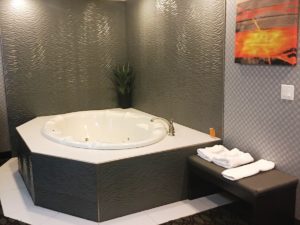 Jacuzzi with bathroom amenities and potted plant, wall mounted art, bench with towels and bath mat, tiled surround
