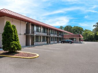 Two story building with covered walkways and exterior guest room entrances, landscaping with small trees and grassy areas, parking spaces