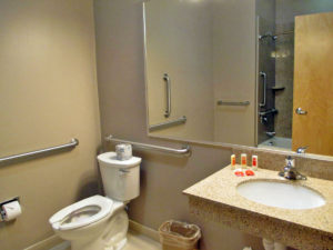 Sink with bathroom amenities, toilet, wall mounted mirror, grab bars, tiled flooring