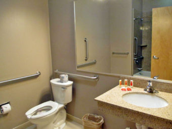 Sink with bathroom amenities, toilet, wall mounted mirror, grab bars, tiled flooring