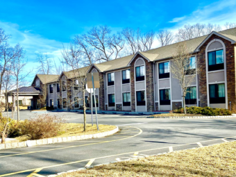 Hotel entrance with drive through canopy, two story building, parking spaces, grassy areas landscaping with small shrubs and trees