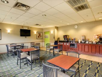 Breakfast area with tables and chairs, breakfast buffet display with juice, coffe and wafle machines, wall mounted TV, wall mounted art, carpet and tile flooring