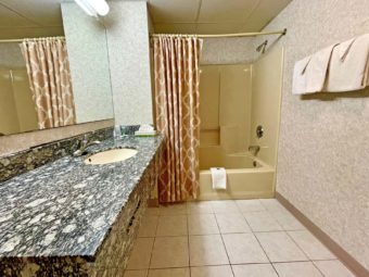 Vanity unit, mirror with overhead lighting, shower tub with shower curtain, towel rail with towels, tiled flooring