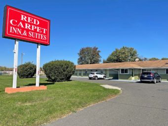 Hotel entrance, branded signage, one story building with exterior room entrances, parking spaces, grassy area with large shrubs