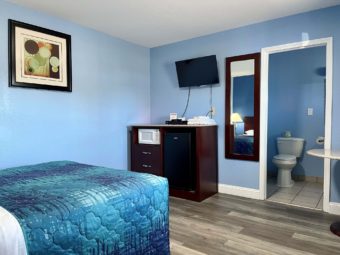 Queen bed, art image, wooden unit with fridge and microwave, wall mounted flatscreen TV, mirror, lamainate flooring, doorway to bathroom, tiled flooring