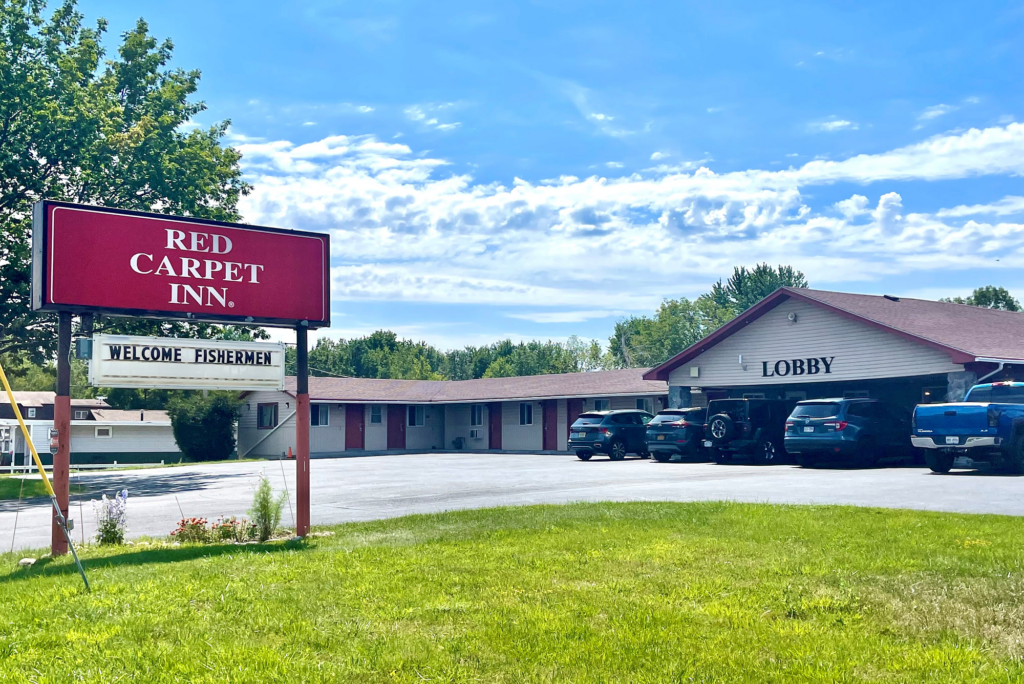 Hotel brand signage, grassy area and flowering shrubs, hotel entrance, one story building with exterior room entrances, parking spaces.