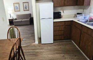 Table and chairs, fridge, microwave, kitchen cabinets, sink, archway to sitting area with sofa, drawer unit with lamp, art images, laminate flooring