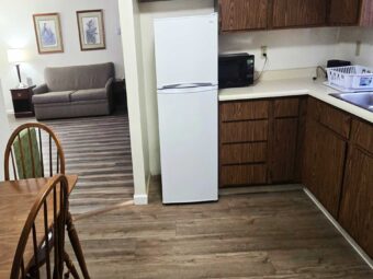 Table and chairs, fridge, microwave, kitchen cabinets, sink, archway to sitting area with sofa, drawer unit with lamp, art images, laminate flooring