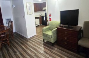 Table and chairs, easy chairs, wooden drawer unit with TV, archway to kitchen area with dishwasher, coffee maker and stove, laminate flooring