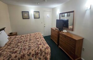 King bed, night stand with clock, art images, wooden drawer units with TV and telephone, mirror, carpet flooring