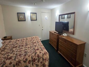 King bed, night stand with clock, art images, wooden drawer units with TV and telephone, mirror, carpet flooring