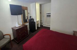 Queen bed, chair, wall mounted TV, wooden drawer unit, mirror, fridge with microwave and coffeemaker, alcove with hanging rail and hangers, portable folding luggage rack, carpet flooring