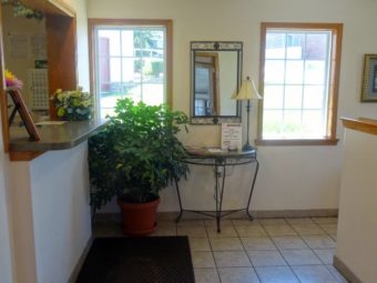 Guest check in desk, potted plant, wall mounted mirror, occasional table with lamp and guest information, tiled flooring