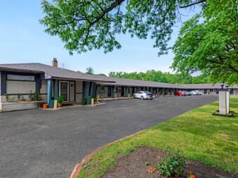 Hotel entrance, one story building with exterior guest room entrances, parking spaces, grassy area with trees