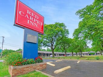 Brand signage, one story building with exterior guest room entrances, parking spaces, grassy area with trees