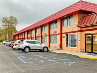 Hotel entrance, two story building, parking spaces, trees and grassy area at end of parking lot