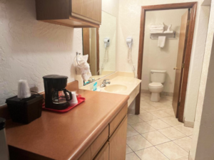 Cupboard with coffeemaker, vanity unit, hairdryer, doorway to bathroom with toilet, towel shelf and rail with towels, tiled flooring