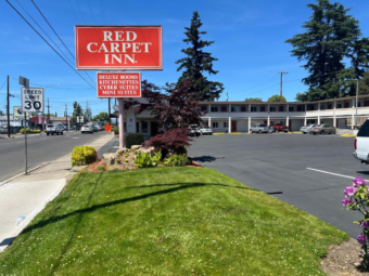 Brand signage, two story building with exterior room entrances and covered walkways, parking spaces, grassy area, landscaping with small shrubs and trees