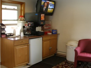 Breakfast counter display with coffee pot, breakfast pastries display case, wall mounted tv, fridge, easy chair, carpet and tiled flooring