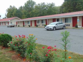 Two single story buildings with exterior room entrances and aprking spaces, grassy area with landscaping flowering bushes and trees