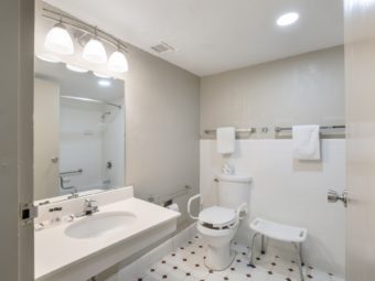 vanity unit, mirror with overhead lights, toilet with grab handles, shower stool, towel rails with towels, tiled flooring