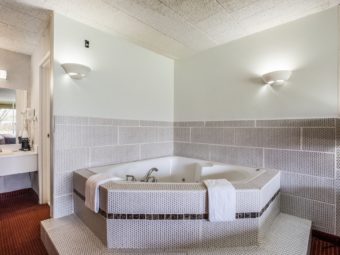 Jacuzzi tub with tiled surround, vanity unit, hairdryer, coffee maker, mirror with overhead light, doorway to bathroom