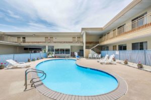 Exterior pool with concrete poolside, pool loungers, ADA pool lift, flowering plants in containers, pool safety fence,two story building with exterior walkways, stairs and room entrances