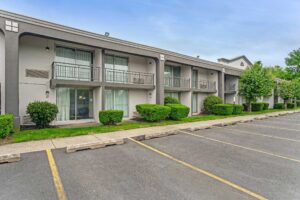 Two story building, balconies with sliding glass doors, landscaping with grassy areas and small bushes, parking spaces