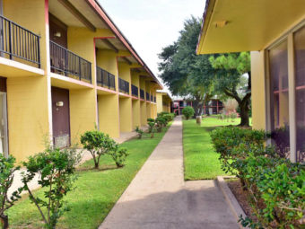 Two story building with balconies on upper level, concrete path, grassy areas with small bushes and trees