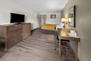 King bed, night stand with telephone, wall bedside lamps, desk with chair, mirror, easy chair, floor lamp, hanging rail with hangers, ironing board and iron, wooden unit with TV, doorway to bathroom, laminate flooring