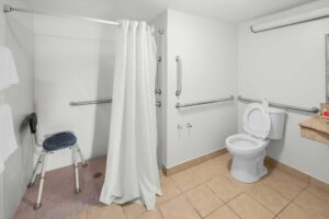 Walk in/roll in shower with chair, grab rails and shower curtain, toilet with grab rails, towel rail with towels, vanity unit, tiled flooring