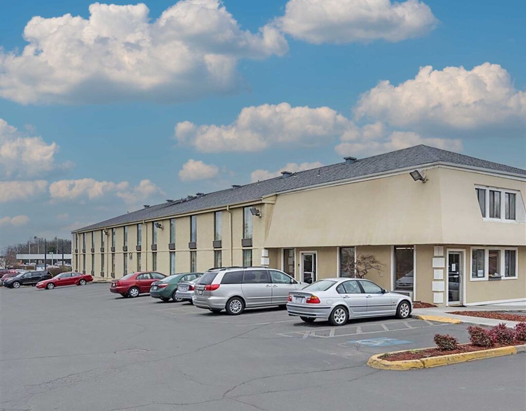 Hotel entrance, two story building, landscaping with small bushes, parking spaces