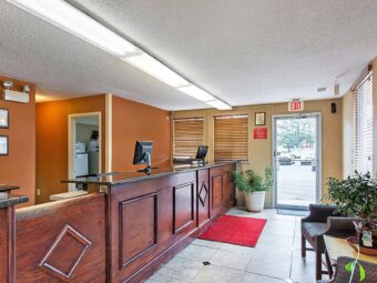 Hotel entrance door, guest check in desk, easy chairs, occasional table, potted plants, tiled flooring with mat