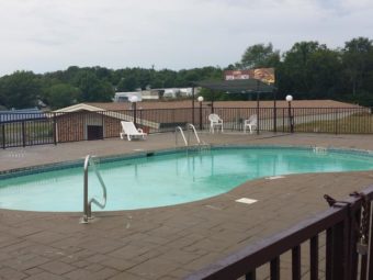 Outdoor pool overlooking hotel, patio furniture on concrete pool surround, metal safety fencing, large trees in the background