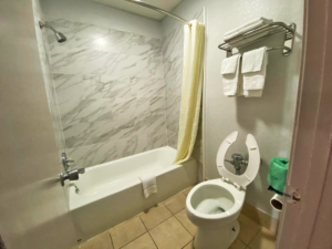 Shower tub with bath mat and shower curtain, toilet, shelf with hanging rail and towels, tiled flooring