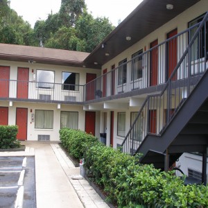 Two story building with exterior walkways, stair and entrances to rooms, plantd areas with small bushes, parking spaces