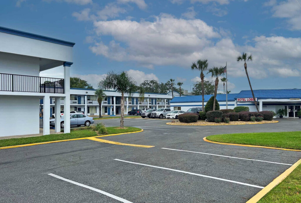 Hotel entrance, two story building with covered walkways, exterior guest room entrances, landscaping with trees, small bushes and grassy areas, parking spaces.