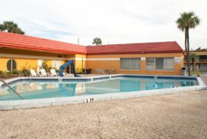 Outside pool, concrete poolside with ada pool chair, plastic pool loungers, small plants and palm tree one story building in the background