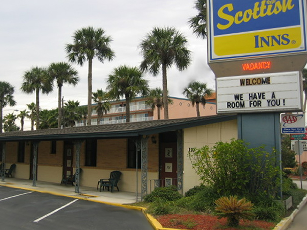 Brand signage, one story building with exterior room entrances, patio chairs, flower bed with small shrubs, palm trees overlooking the hotel