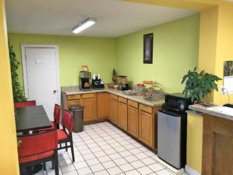 Breakfast counter display with coffe machine, cereal dispensers, fresh fruit display, fridge with microwave, tables and chairs, tiled flooring