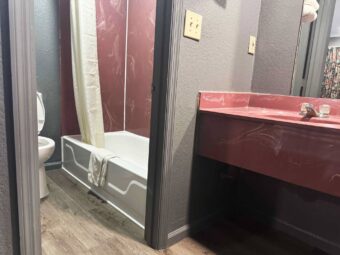 Vanity unit with mirror, doorway leading to shower tub with shower curtain and bath mat, toilet, laminate flooring