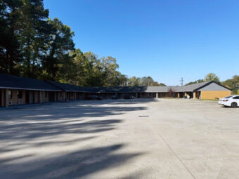one story building with exterior entrances to guest rooms, parking spaces overlooked by tall trees.