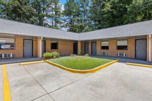 One story building with exterior guest room entrances and covered walkway, parking spaces, grassy area with small shrubs