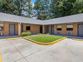 One story building with exterior guest room entrances and covered walkway, parking spaces, grassy area with small shrubs